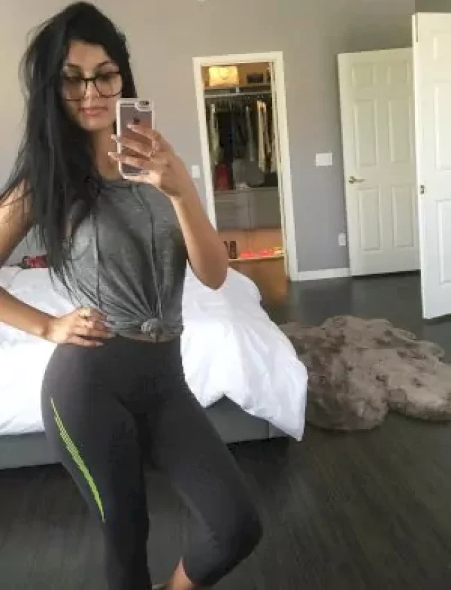 אישה מצטלמת סלפי בחדר שינה במסך מלא, לובשת חולצה אפורה ומכנסיים שחורים עם פס ירוק, ומשקפי ראייה."