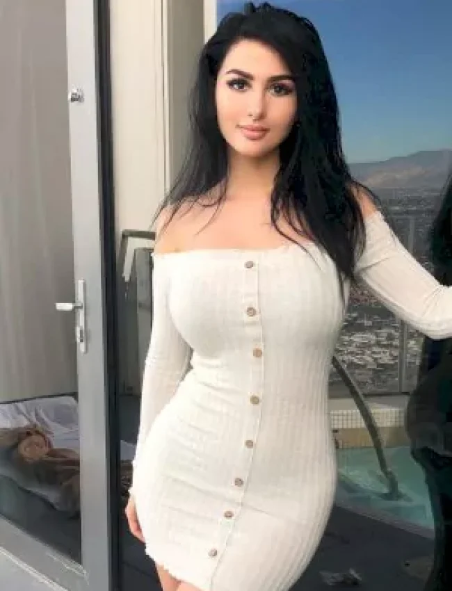 אישה עם שיער כהה לובשת שמלת כפתורים צמודה בצבע לבן ועומדת ליד חלון המראה נוף עירוני.