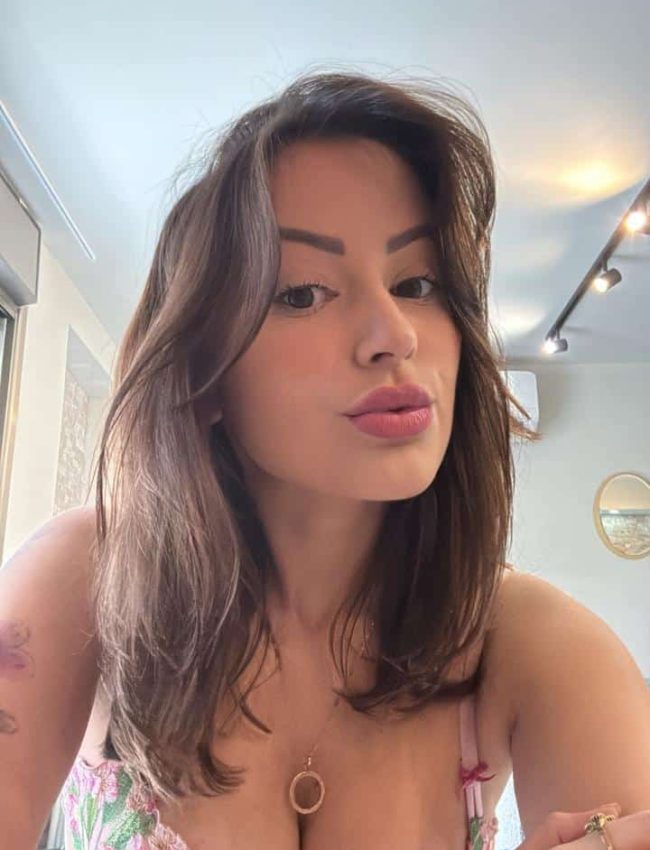 אישה עם שיער חום ארוך קלה עומדת בפנים ונותנת נשיקת שפתי ברווז למצלמה.