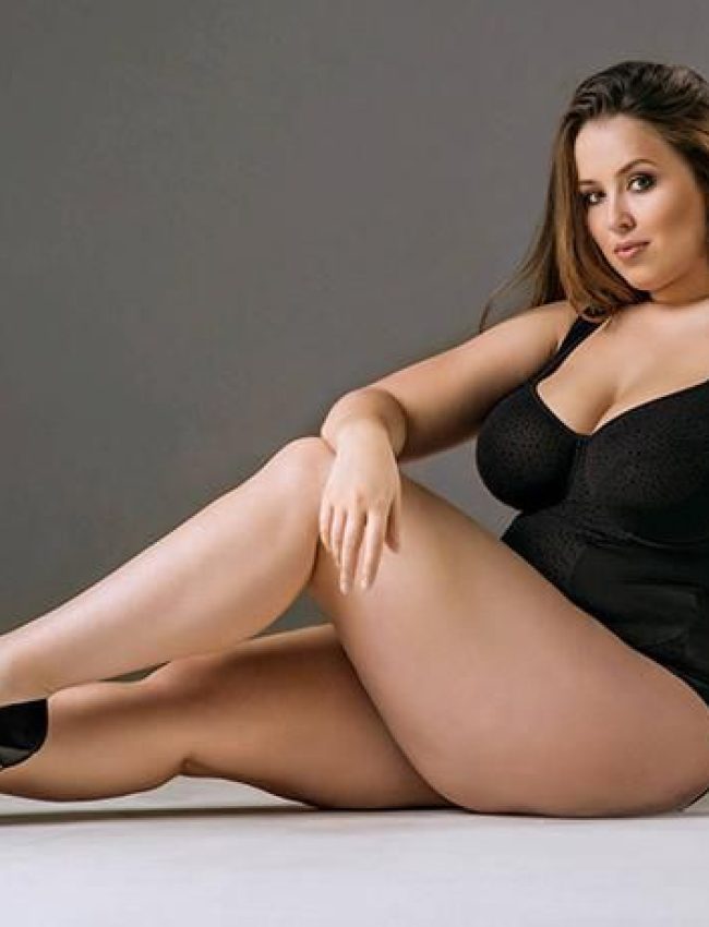 אשה יושבת על הרצפה עם רגליים משוכלות, לובשת בגד גוף שחור ונועלת עקבים שחורים.